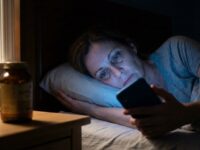 Em um quarto escuro à noite, uma mulher de meia-idade está deitada na cama, acordada, segurando um smartphone cujo brilho azul intenso ilumina seu rosto cansado e seus olhos abertos. Ela olha para a tela com uma expressão de frustração e fadiga. No criado-mudo ao lado, um relógio digital marca 3:17 AM e um frasco âmbar de suplementos está fora de foco.
